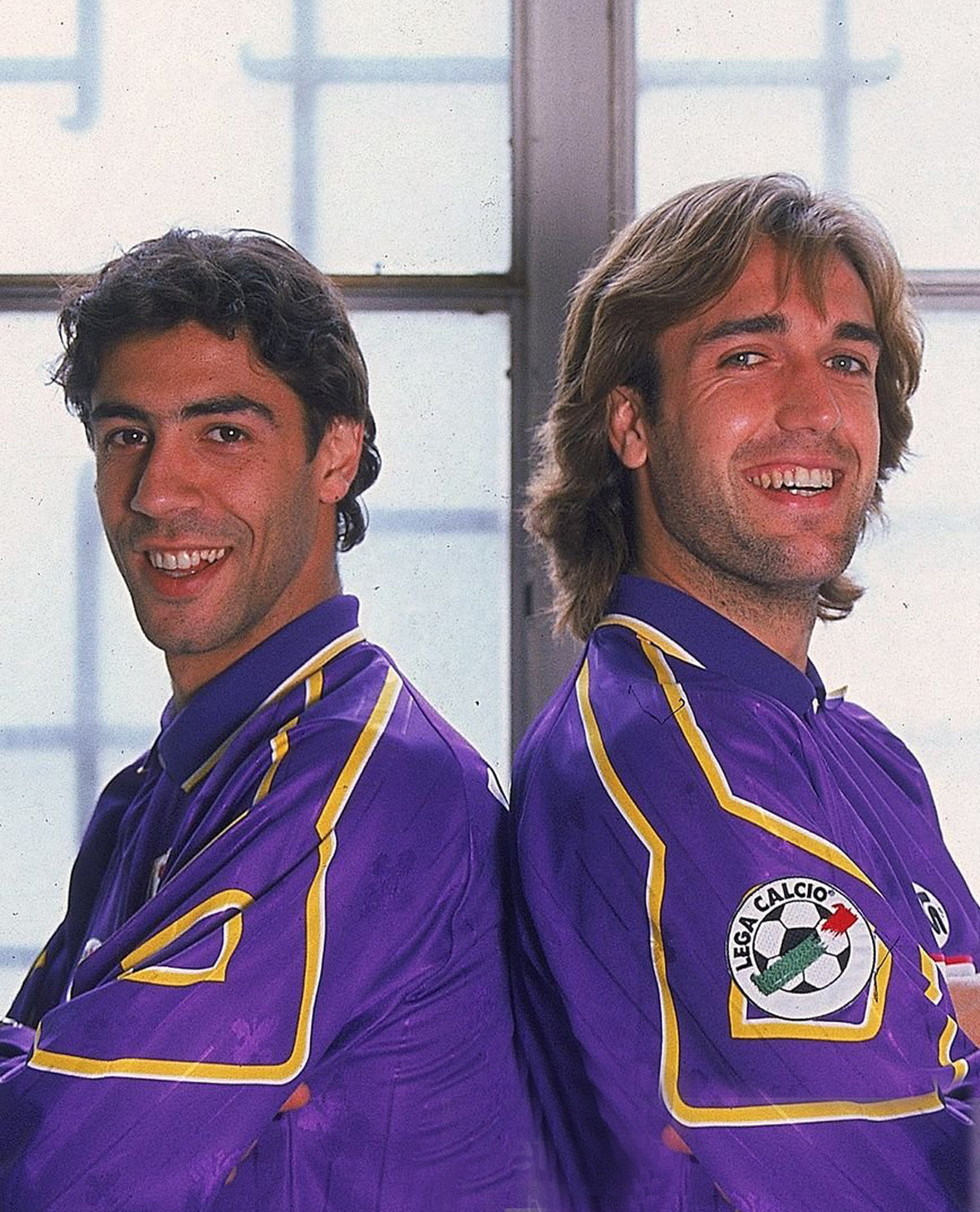 Two men stand back to back, smiling at the camera. They are both wearing electric purple sports uniforms, standing indoors against a window. The man on the left has short, dark brown hair and brown eyes and the man on the right has longer light brown hair and blue eyes.